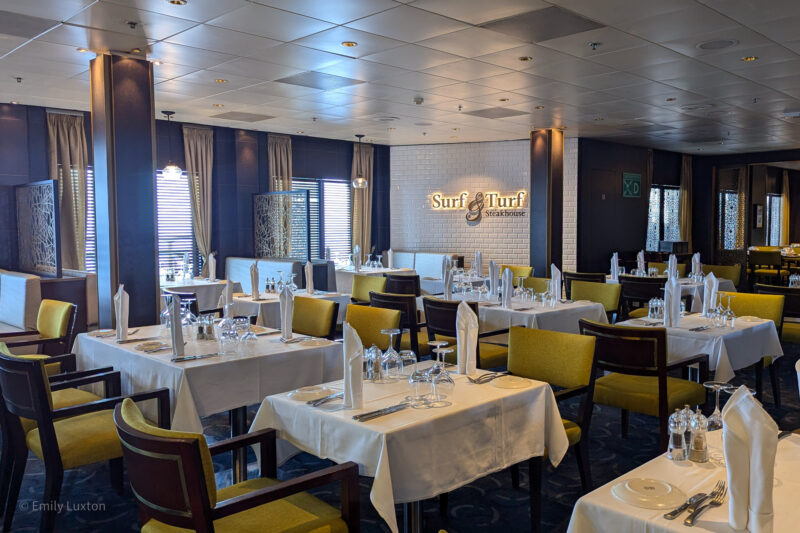 The interior of a restaurant named "Surf & Turf Steakhouse." The restaurant features a modern and elegant design with neatly arranged tables covered with white tablecloths. The walls are decorated with white tiles and dark wood panels, and there are large windows with blinds and curtains allowing natural light to enter.