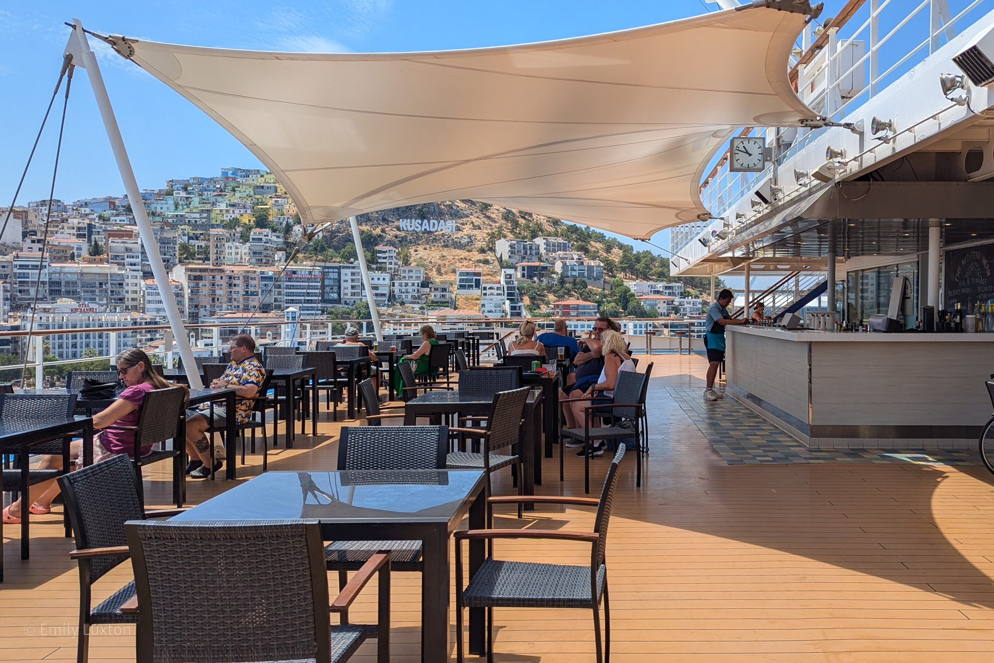 Outdoor seating area on the deck of a cruise ship, with tables and chairs arranged under a large white canopy. In the background, a hillside with numerous buildings prominently displays the word "KUSADASI". To the right, there's a bar area where a person is standing, and several people are seated at the tables.