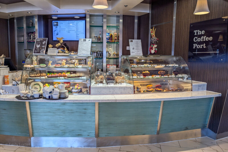 the interior of a coffee shop named "The Coffee Port." The main focus is on the display counter, which is filled with a variety of pastries, desserts, and other baked goods. Behind the counter, there are shelves with various items, including coffee equipment and decorative pieces.