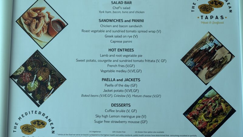 A menu from "The Mediterranean Tapas" restaurant offering a variety of dishes. Categories include Salad Bar, Sandwiches and Panini, Hot Entrees, Paella and Jackets, and Desserts. There are vegetarian (V), vegan (VE), and gluten-free (GF) options.