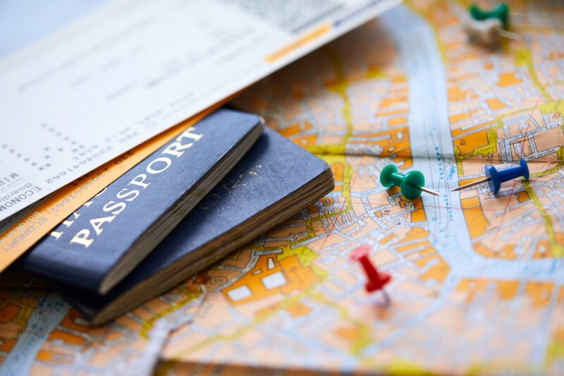 A close-up image shows a blue passport partially covered by a boarding pass, placed on a detailed city map with several push pins in different colors.