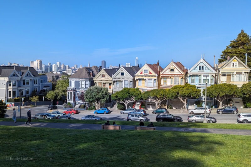 A row of colorful Victorian-style houses, known as the Painted Ladies, in San Francisco, with cars parked on the street and trees in front and a cityscape behind.