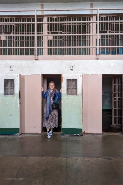 Emily standing in an open cell doorway in an old prison block with pink walls, barred windows and a concrete floor. She is wearing a long leopard print skirt and a denim jacket with a blue scarf.