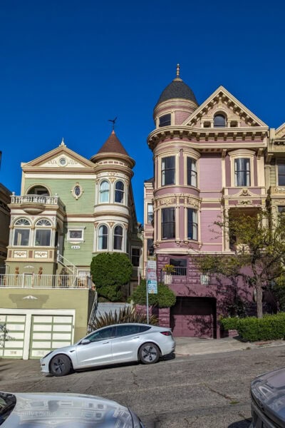 Two adjacent Victorian-style houses with intricate architectural details, painted in light green and light purple, with ornate trims. A white Tesla is parked on a steep street in front.