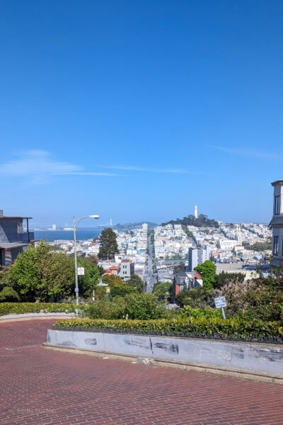 A view from the top of Lombard Street in San Francisco, showing the steep, brick-paved road with eight hairpin turns, residential buildings, and the San Francisco cityscape with the Coit Tower and Bay Bridge in the background.