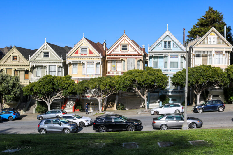 A row of colorful Victorian-style houses, known as the Painted Ladies, with cars parked on the street and trees in front. Two Days in San Francisco.