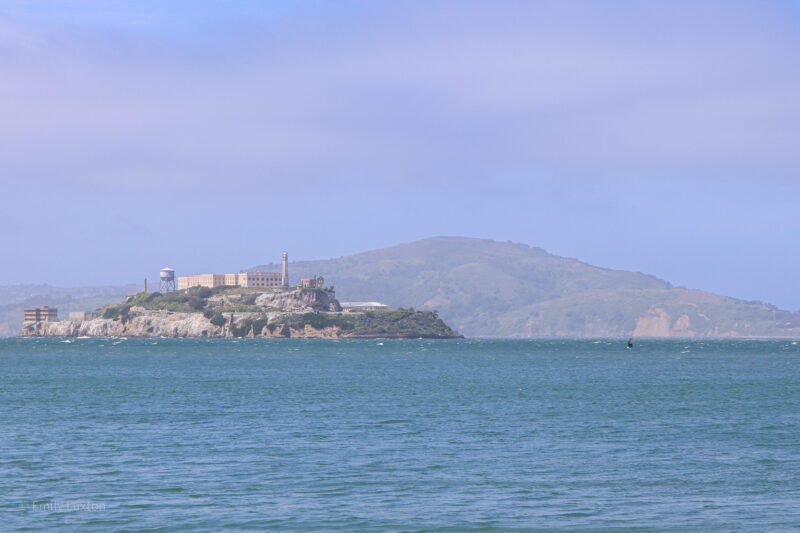 View of Alcatraz, a small island surrounded by sea with a larger hilly landmass in the distance behind it. The island has several old prison buildings on it. 