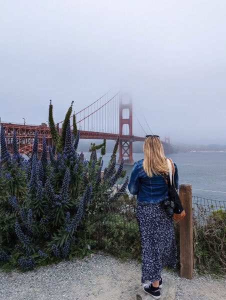 Emily wearing a long blue dress and blue denim jacket standing on a gravel path, facing away from the camera, looking toward the Golden Gate Bridge, which is partially shrouded in fog.