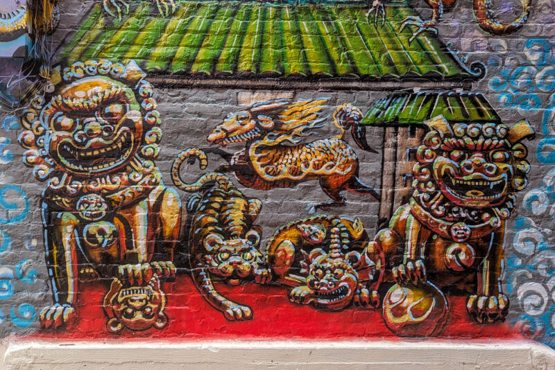 A colorful mural on a brick wall depicting Chinese mythical creatures, including two ornate lion-like figures, a tiger, and a dragon-like creature.