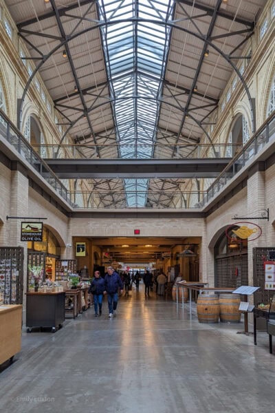 Interior of the Ferry Building in San Francisco with a high curved glass ceiling. The walkway is lined with small shops and has a grey tiled floor.