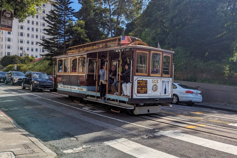 A classic San Francisco cable car on the Powell & Hyde line, with passengers visible. The cable car is brown and cream-colored, displaying the number 23. A white building, trees, and parked cars are in the background.