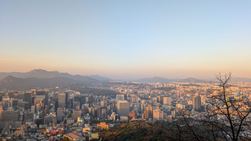 A panoramic view of Seoul cityscape at sunset, featuring a mix of modern skyscrapers and smaller buildings. In the background, mountain ranges are visible under a clear sky with a warm, golden hue. A leafless tree stands on the right side of the image, silhouetted against the setting sun. Lead image for the post: Is South Korea Safe for Solo Female Travellers?