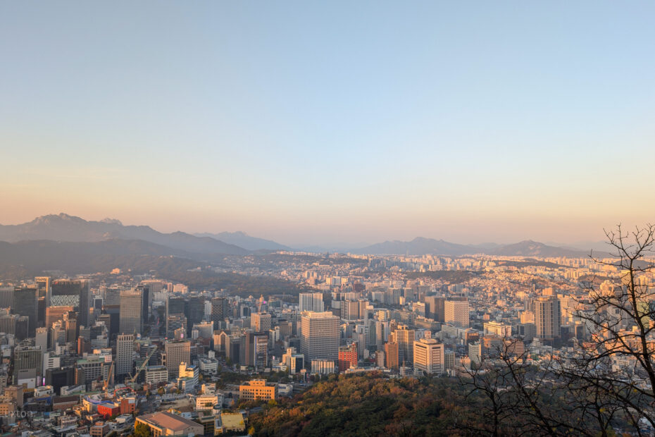 A panoramic view of a cityscape at sunset, featuring a mix of modern skyscrapers and smaller buildings. In the background, mountain ranges are visible under a clear sky with a warm, golden hue. A leafless tree stands on the right side of the image, silhouetted against the setting sun.