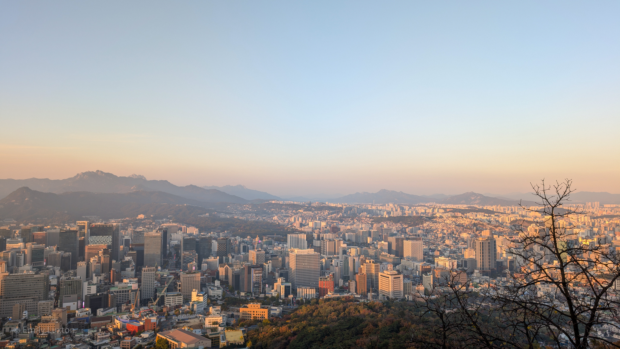 A panoramic view of a cityscape at sunset, featuring a mix of modern skyscrapers and smaller buildings. In the background, mountain ranges are visible under a clear sky with a warm, golden hue. A leafless tree stands on the right side of the image, silhouetted against the setting sun.