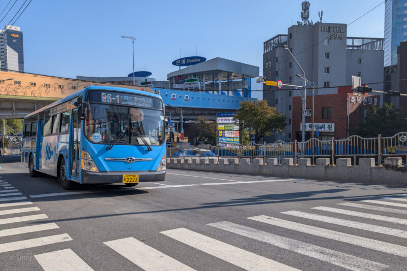 A blue bus with the number 99 on its display is driving through a crosswalk in an urban area. The bus has a yellow license plate with the number 2223. In the background, there are various buildings, including one labeled "Hotel 99.9" and another with the sign "Cheonma." An overpass with animal drawings and a blue structure is also visible.