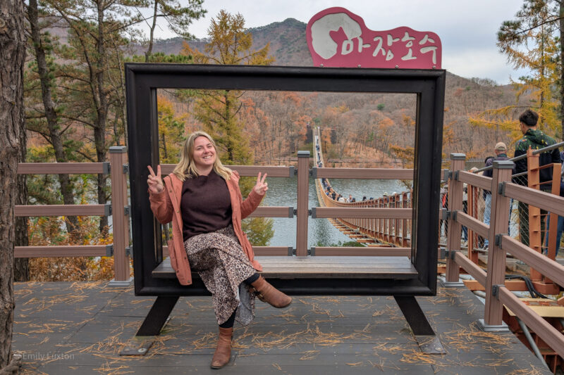 Emily is sitting on a bench framed by a large rectangular structure, making a peace sign with both hands. Behind them, a suspension bridge spans a body of water, leading into a forest with autumn-colored trees. Several people are walking on the bridge. A sign in Korean reads "Majang Lake." Emily is wearing a leopard print skirt, brown leather boots, a burgundy jumper and a long peach-orange raincoat.