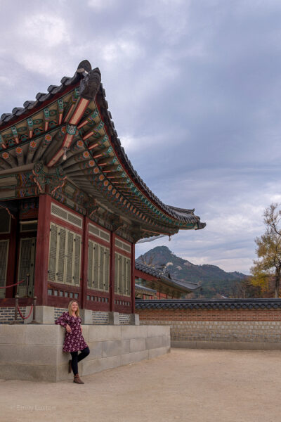 A traditional Korean building elevated on a stone platform features colorful eaves and ornate patterns. Emily is leaning against the platform. In the background, scenic mountains and a cloudy sky add to the picturesque beauty. Emily is travelling in South Korea solo, wearing a dark red dress with pink floral print, black leggings, and brown leather boots.