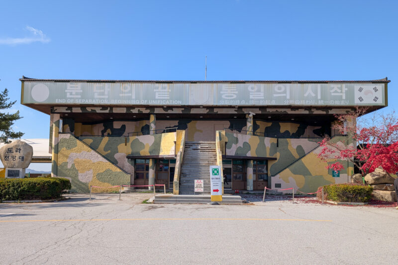 A building with a camouflage pattern on its exterior walls features a staircase leading up to the entrance. Signs in English, Korean, Chinese, and Japanese read "END OF SEPARATION, BEGINNING OF UNIFICATION." A stone monument with Korean text stands to the left. Trees and bushes surround the building, with one tree having red leaves.