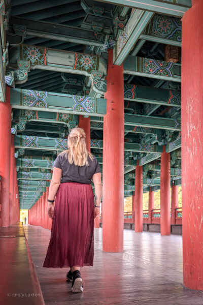 Emily is walking alone through a traditional East Asian architectural structure with red columns and intricately decorated beams and ceiling. The individual is dressed in a black top, long maroon skirt, and black shoes. The vibrant colors and detailed patterns of the structure are visually captivating, drawing the viewer's eye towards the vanishing point in the distance. Solo female travel safety in South Korea.