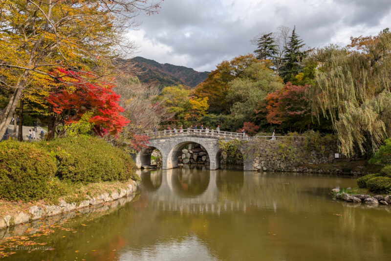 A scenic view of a stone bridge over a calm body of water, surrounded by autumn foliage. The bridge has multiple arches and a railing, with people walking across it. The trees around the bridge display vibrant fall colors, including shades of red, yellow, and green. The reflection of the bridge and trees can be seen in the water below. In the background, there are hills or mountains under a partly cloudy sky.