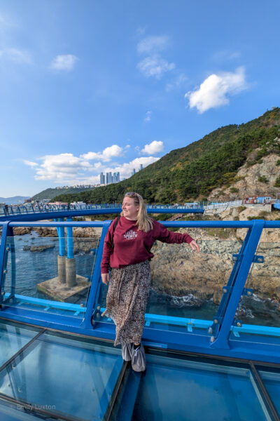 Emily is standing on a glass-floored observation deck with a scenic view of the ocean and rocky coastline. She is wearing a maroon sweatshirt with the text "CLUB SPORTIF" on it, a long leopard print skirt, and gray shoe covers. The background features a lush green hillside and a distant cityscape with tall buildings under a bright blue sky with scattered clouds.