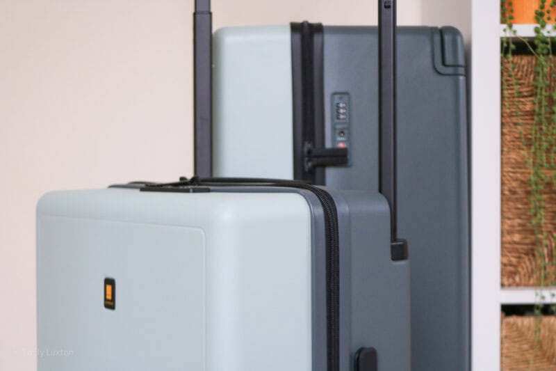 A light blue luggage with a black zipper and a small orange and black logo, standing in front of a dark gray luggage with a combination lock.