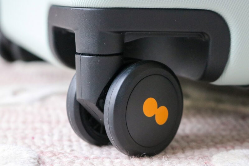 Close-up view of a black caster wheel attached to light-colored luggage with a textured finish. The wheel has an orange logo resembling two connected dots.