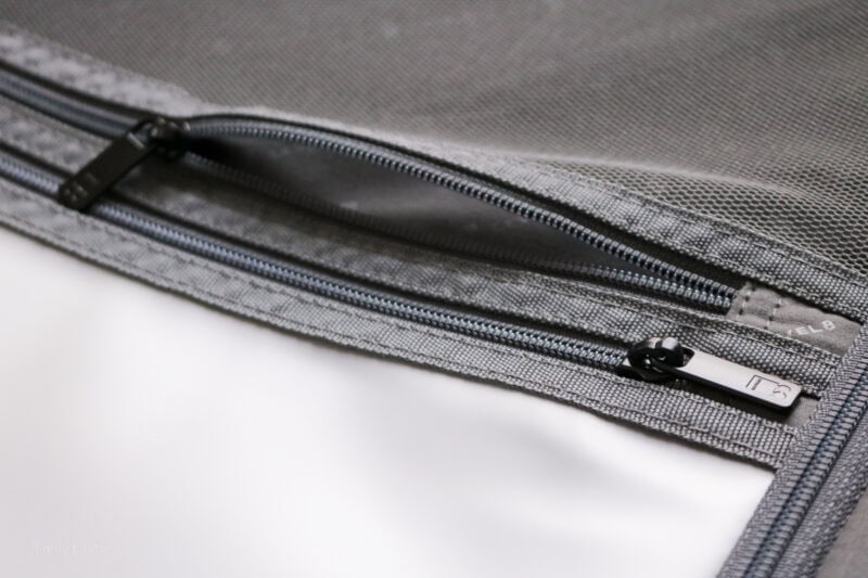 Close-up view of a gray fabric item with two zippers. The zippers are parallel to each other, with one partially open, revealing the interior. The metallic zipper pulls include a small logo or text. The fabric appears durable and textured.