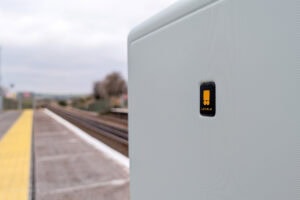 Close-up view of a textured surface, likely a piece of luggage, featuring a small black and yellow logo with the word "LEVEL8." In the blurred background, there's a train station platform with yellow tactile paving and train tracks extending into the distance.