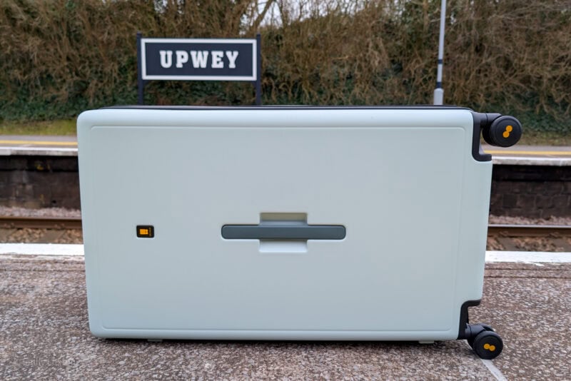 Light-colored suitcase lying on its side on a train station platform. The suitcase has four wheels, with yellow accents and a small rectangular indicator. In the background, there is a sign that reads "UPWEY," with platform and train tracks visible.