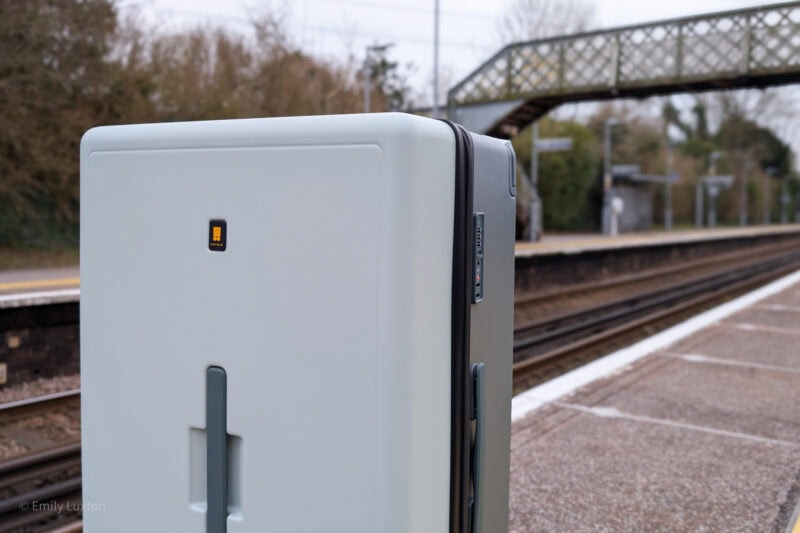 Close-up view of a light gray suitcase with a black handle and four wheels, standing on a train platform. The suitcase features a small rectangular logo with an orange icon. There is a bridge and train tracks in the background.