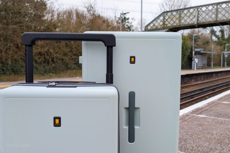 Two light blue pieces of luggage with black handles and LEVEL8 logos, standing on a train station platform with tracks and a pedestrian bridge in the background.