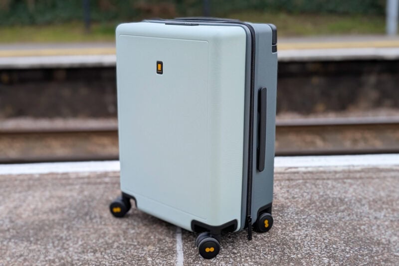 Light-colored, hard-shell suitcase with four wheels on a platform, possibly at a train station. The background shows blurred train tracks and platform edge.