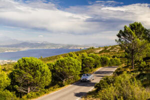 Convertible car on a winding road in Corsica France, surrounded by lush green trees, with a lake and mountains in the distance under a partly cloudy sky.