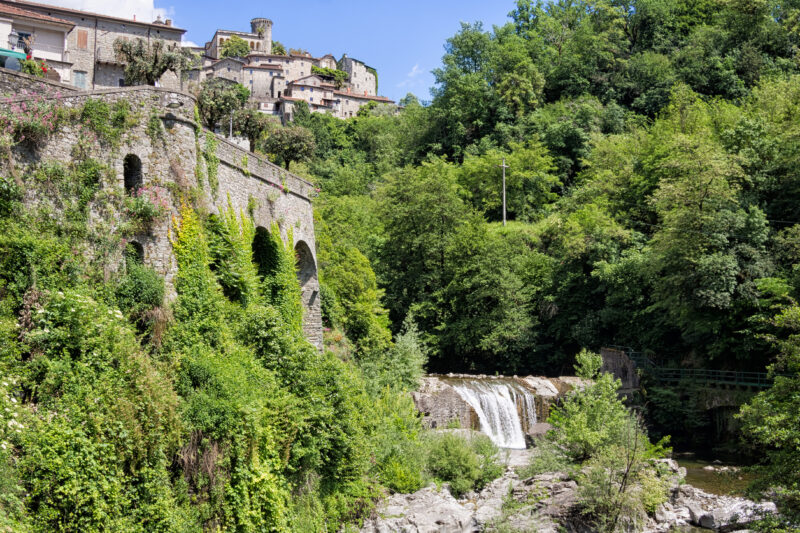 Bagnone town with grey stone buildings on a hillside surrounded by trees next to a river and waterfall. 