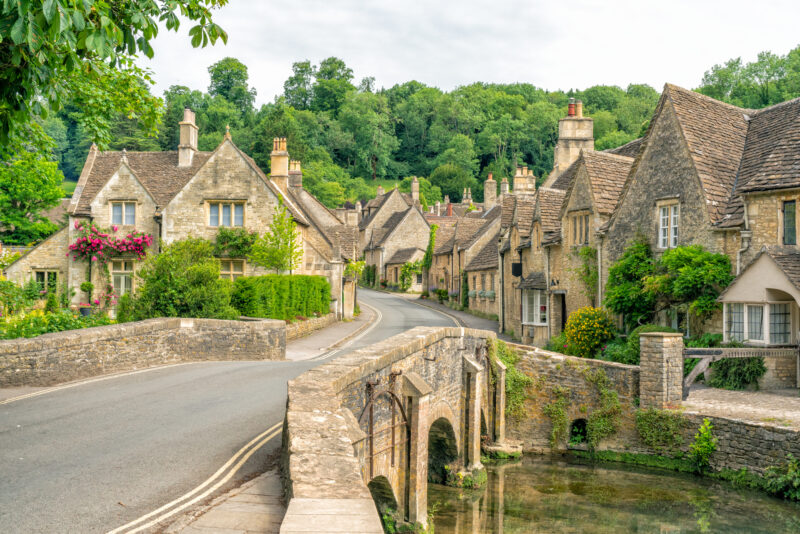 stone bridge over a river with a small village of beige stone cottages and grey slate roofs surrounded by green leafy treets