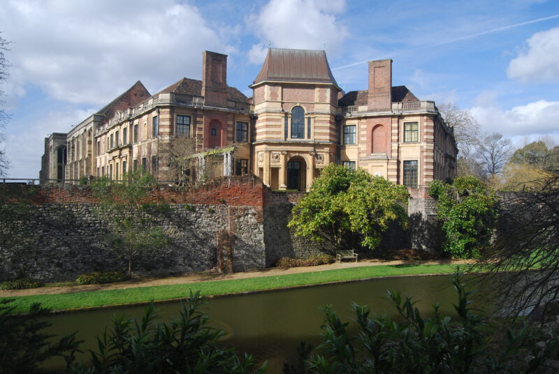 Exterior of a large red brick mansion with a grey slate roof surrounded by leafy trees on a sunny day with blue sky. 