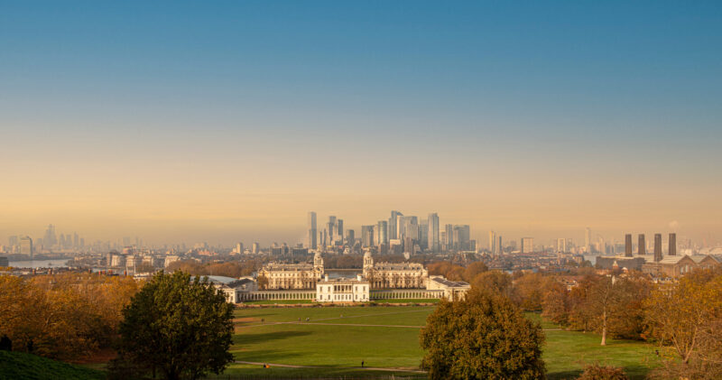 View across a large green lawn in Greenwich park framed by trees with a white mansion in the distance and behind it London city skyline with numerous skyscrapers against a dusky sky