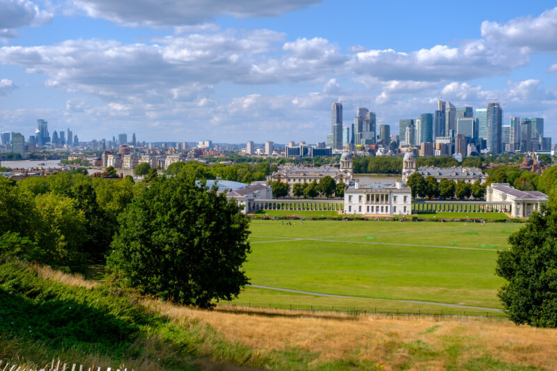 View from a small grassy hill in Greenwich Park with a large lawn framed by trees. On the far side is a large white mansion and behind that the London city skyline with many skyscrapers. 