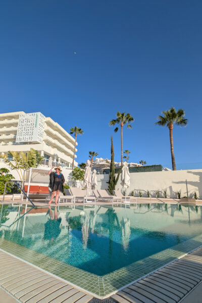 Outdoor pool area with a white building labeled 'Iberostar,' palm trees, and sun lougners. Emily is on the other side of te pool wearing a black cover up and a wide brimmed floppy sunhat.