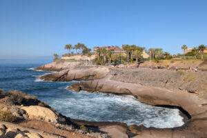 Rocky cliffs with ocean waves, a building with red-tiled roofs, palm trees, and a clear blue sky, people walking on a pathway