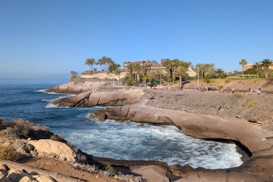 Rocky cliffs with ocean waves, a building with red-tiled roofs, palm trees, and a clear blue sky, people walking on a pathway