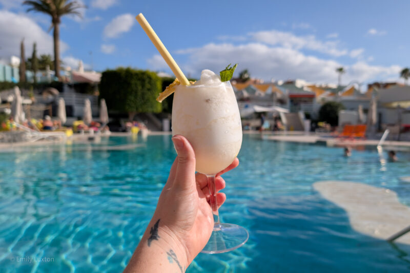 Hand holding a white creamy cocktail with a yellow straw in front of a pool, with lounge chairs, umbrellas, and palm trees in the background