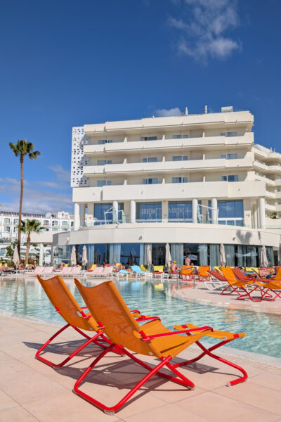 Modern hotel with multiple floors and balconies, a pool surrounded by sun loungers, and people relaxing under clear blue skies with two bright orange deckchairs in the foreground.