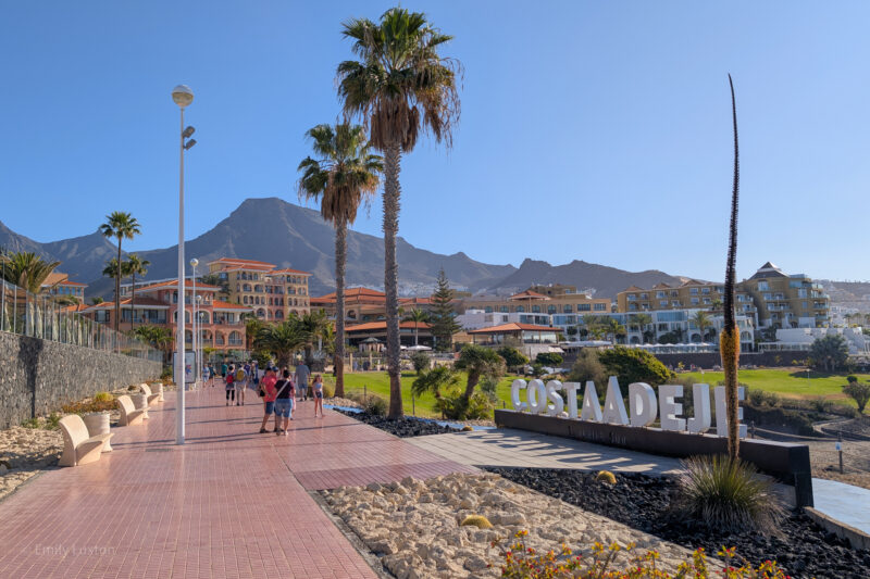 Tiled walkway in Tenerife with palm trees, benches, people walking, a sign reading 'COSTA ADEJE,' and buildings with mountain views.