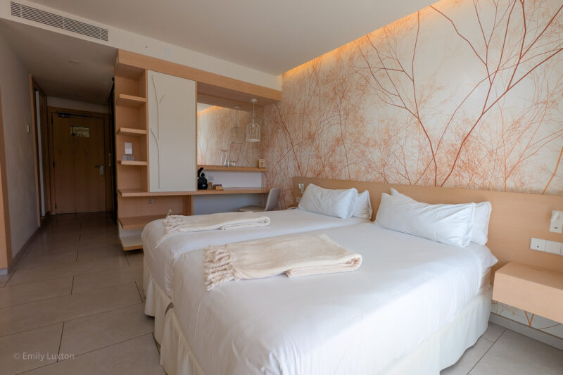 Modern hotel room with two twin beds, beige blankets, nature-inspired wall design, a desk with a coffee machine, and a wardrobe with open shelves.