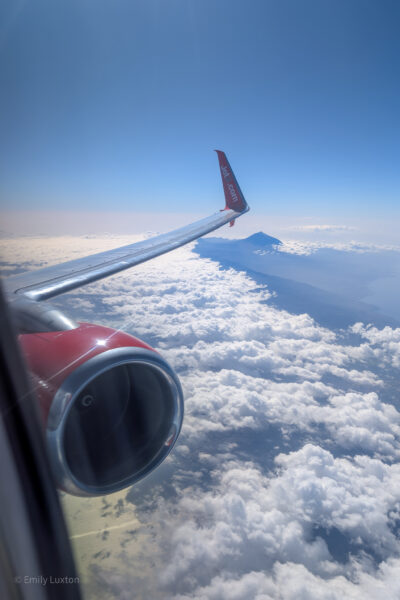 Airplane wing with 'Jet2.com' logo and engine, seen through a window. Vast clouds below and a clear blue sky, with a mountain peak in the distance.