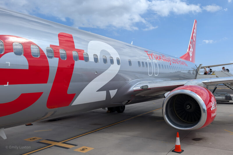 Jet2.com airplane parked on the tarmac, silver with red and white branding, engine visible with traffic cone nearby, people in the background. Jet2 holidays flight to Tenerife review.