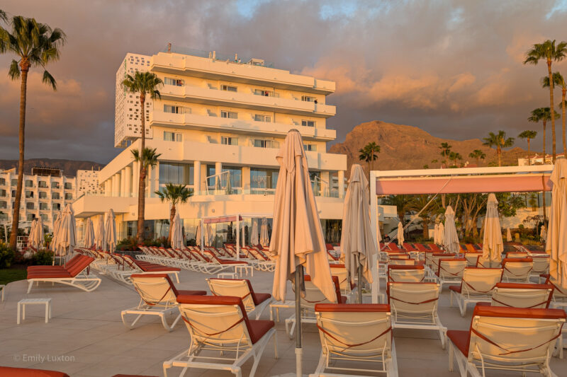 Resort pool area during sunset with lounge chairs, umbrellas, palm trees, multi-story building with balconies, and mountains in the background.