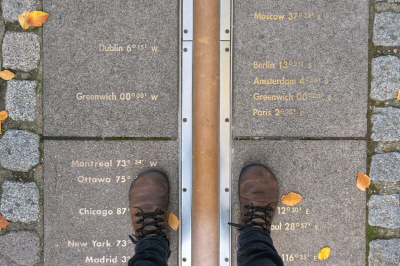 Feet straddling the Prime Meridian at Greenwich, London, with longitude markers engraved on the ground, representing global time zones.